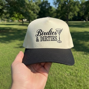 May include: A cream-colored trucker hat with a black brim. The hat features the text "Birdies & Dirties" with a martini glass graphic in black. The hat is held up in front of a green background.