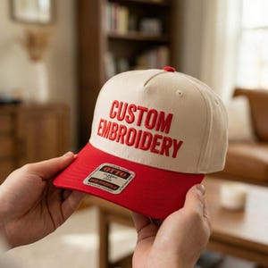 May include: A beige and red baseball cap with the words "CUSTOM EMBROIDERY" embroidered in red. The cap has a red brim and a small tag that reads "OTTO CLASSIC CFM MID PROFILE".