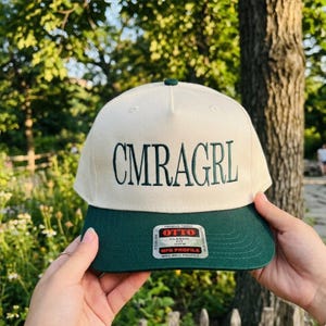 May include: A cream-colored baseball cap with a dark green brim. The text "CMRAGRL" is embroidered in dark green on the front. The cap has a green button on top and a "OTTO" label on the brim.
