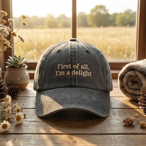 May include: A gray baseball cap with the embroidered text "First of all, I'm a delight." The hat is made of a soft, worn material. The cap is sitting on a wooden surface with a window in the background.
