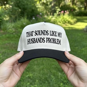 May include: A cream-colored baseball cap with a black brim. The cap features the phrase "THAT SOUNDS LIKE MY HUSBANDS PROBLEM" in bold, black lettering. The hat is held up against a blurred green background.