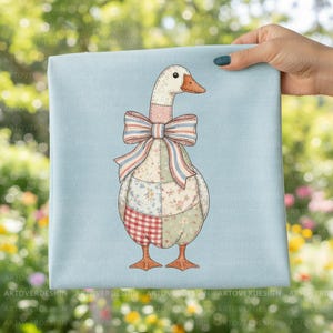 Könnte beinhalten: Ein hellblaues quadratisches Tuch mit einer skurrilen Illustration einer Gans. Die Gans ist mit einem Patchwork-Design mit Blumen- und Vichy-Mustern und einer großen Schleife verziert. Die Gans hat orangefarbene Füße und einen Schnabel.