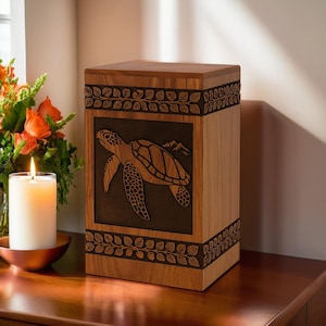 May include: Wooden urn with a carved sea turtle design. The rectangular urn is made of light-coloured wood with a dark brown turtle and mountain scene. Leaf patterns border the top and bottom.