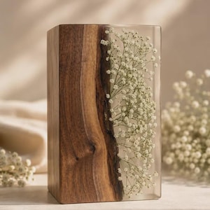 Op de afbeelding: Een rechthoekig blok met een verticaal gedeelte van natuurlijk bruin hout met een zichtbare houtnerf, verbonden met een helder harsgedeelte waarin witte gipskruidbloemen zijn verwerkt. Het object staat rechtop met een gladde afwerking.