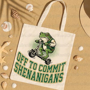 K&ouml;nnte beinhalten: Beige Tragetasche mit einem Cartoon-Frosch auf einem Roller. Der Frosch ist gr&uuml;n und tr&auml;gt eine gr&uuml;n-wei&szlig;e Jacke. Der Text "OFF TO COMMIT SHENANIGANS" ist gr&uuml;n auf der Tasche aufgedruckt.