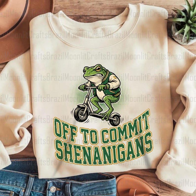 K&ouml;nnte beinhalten: Cremefarbenes Sweatshirt mit einem gr&uuml;nen Frosch auf einem Roller. Der Frosch tr&auml;gt eine gr&uuml;ne Jacke und der Text "OFF TO COMMIT SHENANIGANS" steht darunter. Ein lustiges, skurriles Design.