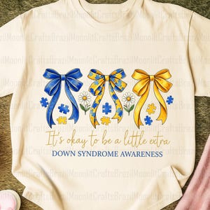 May include: A cream-coloured t-shirt with a design promoting Down Syndrome awareness. The design features three bows in blue, yellow, and a yellow and blue pattern, with puzzle pieces and daisies. Text reads "It's okay to be a little extra" and "DOWN SYNDROME AWARENESS."