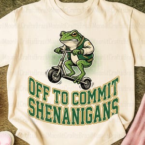 K&ouml;nnte beinhalten: Cremefarbenes T-Shirt mit einem Cartoon-Frosch auf einem Roller, der eine gr&uuml;n-wei&szlig;e Jacke tr&auml;gt. Der Text "OFF TO COMMIT SHENANIGANS" ist in gr&uuml;nen und goldenen Buchstaben gehalten. Ein lustiges, skurriles Design.