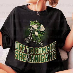 K&ouml;nnte beinhalten: Schwarzes T-Shirt mit einer gr&uuml;nen Froschfigur auf einem Roller, bekleidet mit einer gr&uuml;n-wei&szlig;en Jacke. Der Text "OFF TO COMMIT SHENANIGANS" ist in Gr&uuml;n und Gold gehalten. Ein witziges Design.