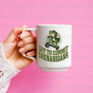K&ouml;nnte beinhalten: Wei&szlig;e Keramiktasse mit einem gr&uuml;nen Frosch auf einem Roller und dem Text "OFF TO COMMIT SHENANIGANS". Die Tasse wird von einer Person gehalten. Der Hintergrund ist rosa.