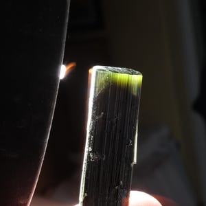 Pu&ograve; includere: Un cristallo di tormalina grezzo e allungato con una consistenza striata verticale. Il minerale mostra un colore verde scuro profondo che sfuma in un verde oliva pi&ugrave; chiaro e traslucido sul bordo superiore dove la luce attraversa la struttura.