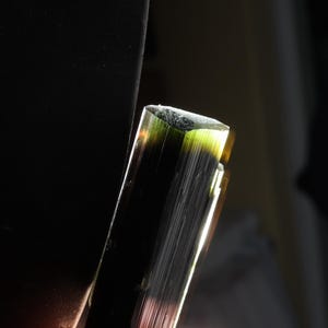 Pu&ograve; includere: Un cristallo di tormalina grezzo e allungato con una consistenza striata verticale. Il minerale mostra una sfumatura di colore che passa da un nero profondo e scuro alla base a un verde oliva vibrante e traslucido sulla sommit&agrave; piatta.