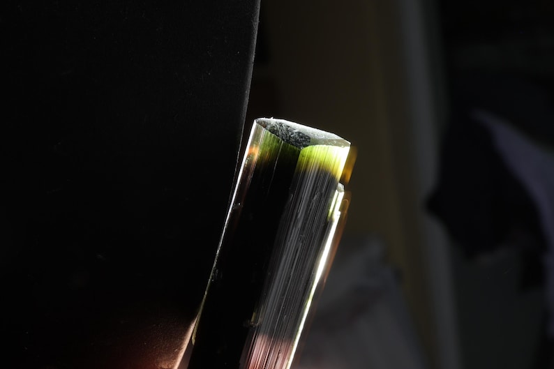Pu&ograve; includere: Un cristallo di tormalina grezzo e allungato con una consistenza striata verticale. Il minerale mostra una sfumatura di colore che passa da un nero profondo e scuro alla base a un verde oliva vibrante e traslucido sulla sommit&agrave; piatta.