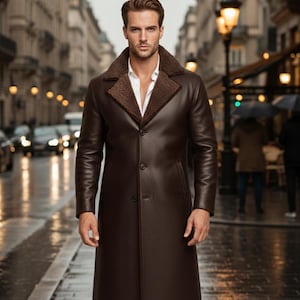 May include: A long, dark brown leather coat with a shearling collar. The coat is buttoned up and worn over a white collared shirt. The image is taken on a city street with blurred lights and buildings in the background.