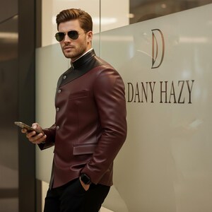 May include: A burgundy leather jacket with a black shoulder panel and a mandarin collar. The jacket has a unique asymmetrical design with button closures and a small pocket. The text "DANY HAZY" is visible on the glass behind the jacket.