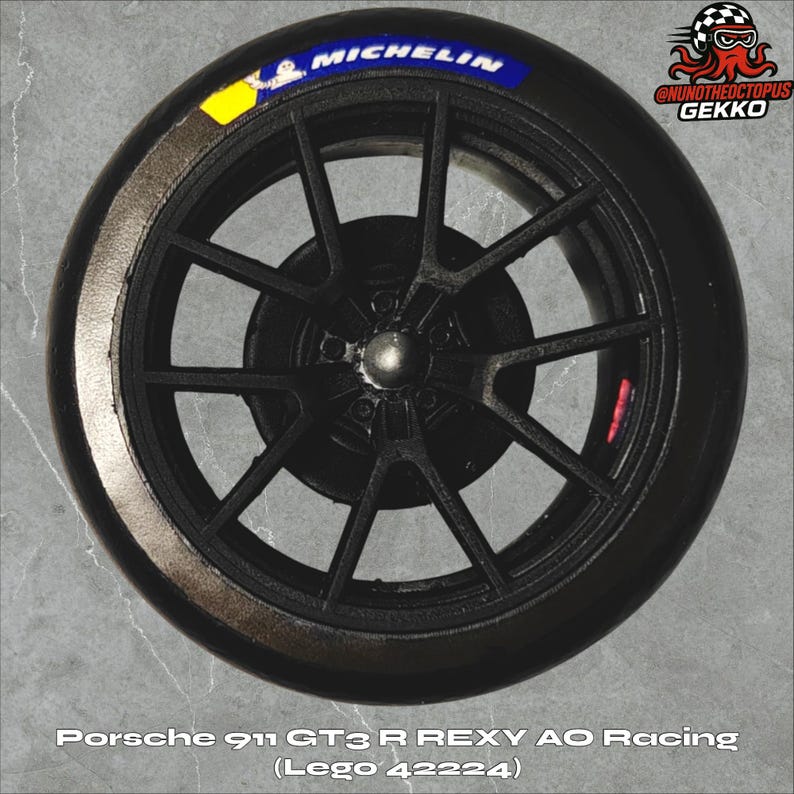 May include: Black racing wheel with a Michelin tire. The wheel has a dark gray spoke design and a silver center cap. The tire has the text "MICHELIN" in blue and white. The text "Porsche 911 GT3 R REXY AO Racing (Lego 42224)" is at the bottom.