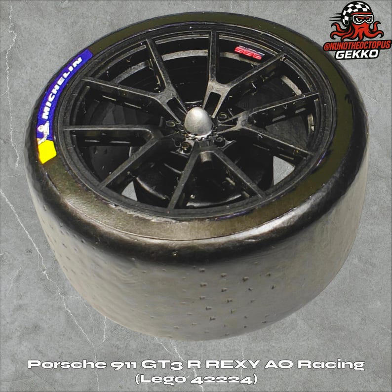 May include: A black racing tire with a black spoke wheel. The tire has a Michelin logo and the text "Porsche 911 GT3 R REXY AO Racing (Lego 42224)". The wheel has a silver center cap.