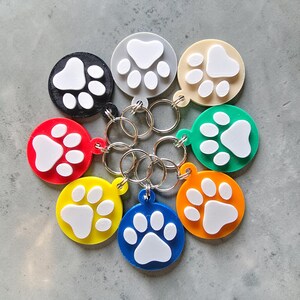 May include: A collection of eight circular keychains in black, grey, beige, green, orange, blue, yellow, and red. Each keychain features a raised white paw print design on the front and includes a silver metal split ring for attachment.