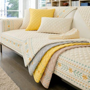 Könnte beinhalten: Ein cremefarbenes Sofa mit mehreren cremefarbenen und gelben Kissen. Das Sofa ist mit einer gesteppten Tagesdecke bedeckt, und gefaltete Decken in Gelb, Grau und Rosa sind über die Seite drapiert. Die Tagesdecke und die Decken haben Blumenstickereien.