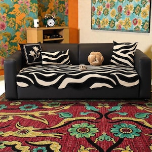 May include: A black leather sofa with a zebra-striped cover and decorative pillows. The pillows feature zebra patterns and floral designs. A decorative rug with a floral pattern is in the foreground. A small wooden table and artwork are in the background.