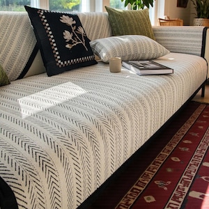 以下が含まれることがあります： 黒のヘリンボーン模様のクリーム色のカバーが付いたモダンなソファ。さまざまなデザインの黒、緑、クリーム色の装飾枕がソファに配置されています。本と小さなセラミックカップがソファの上にあります。