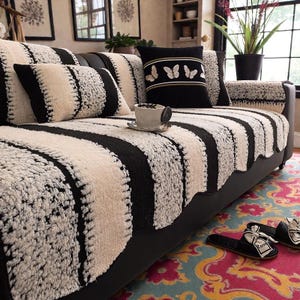 May include: A black sofa with a striped black and cream-colored cover and matching pillows. A black pillow with butterfly embroidery and a teacup on a saucer are on the sofa. Black slippers with a bow are on a colorful rug.