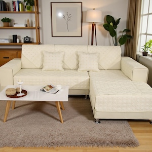 Puede incluir: Un sofá seccional color crema con cojines a juego y una funda acolchada. Una mesa de centro blanca con patas de madera descansa sobre una alfombra marrón peluda. Una lámpara y una obra de arte enmarcada son visibles en el fondo.