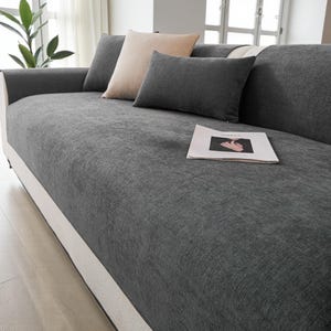May include: A grey sofa with a matching grey cover and several throw pillows in shades of grey and beige. A magazine rests on the sofa. The sofa has a white base.