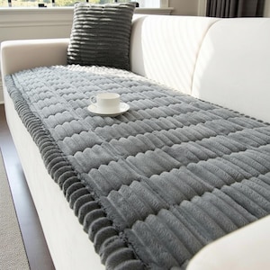 May include: A grey, textured sofa cover with a matching cushion. The cover has a ribbed design and a soft, plush appearance. A white teacup and saucer sit on the cover. The sofa is white.