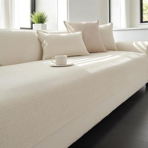 Op de afbeelding: Een crèmekleurige bank met drie decoratieve kussens en een kleine theekop op een schotel. De bank heeft een textuurstof en staat in een goed verlichte kamer. De kussens zijn in crème- en beigetinten.