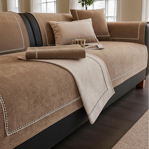 May include: A brown sofa with matching cushions and a throw blanket. The sofa and cushions are a textured fabric with white stitching around the edges. The throw blanket is a lighter shade of brown. A cup and magazine are on the sofa.