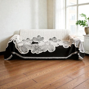 May include: A black and white floral patterned sofa cover draped over a sofa. The cover features large, stylized flower designs with a scalloped edge. The sofa has a striped back and is set against a neutral wall and a window.