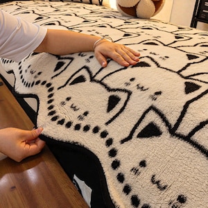 May include: Cream-colored bedspread featuring a repeating pattern of black cat faces. The bedspread has a soft, textured appearance and is being adjusted by a person's hand. The edges are trimmed with black fabric.