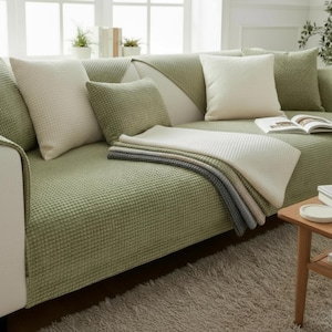 May include: A green textured sofa cover with matching pillows and a folded blanket. The sofa is in a living room setting with a neutral rug and wooden side table. The pillows are in shades of green and cream.