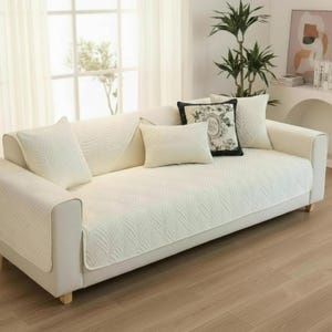 Puede incluir: Un sofá blanco con una funda estampada y varios cojines decorativos. El sofá tiene una estructura de madera clara y está colocado frente a una ventana. Los cojines incluyen un diseño floral en blanco y negro.