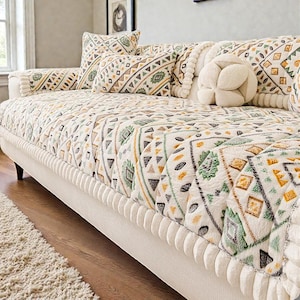 Könnte beinhalten: Ein cremefarbenes Sofa mit einer gemusterten Steppdecke und passenden Kissen. Die Steppdecke zeigt geometrische Muster in Grün, Gelb und Braun. Ein dekoratives, rundes, cremefarbenes Kissen ist ebenfalls vorhanden. Das Sofa steht auf dunklen Holzbeinen.