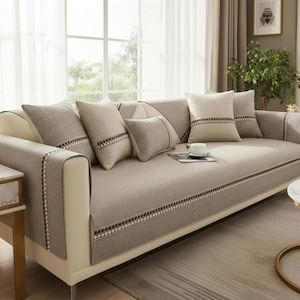 Op de afbeelding: Een beige bank met meerdere sierkussens. De bank is bedekt met een stoffen hoes met textuur en een patroonrand. De kussens zijn in verschillende maten en tinten beige en bruin. De kamer heeft een neutrale kleurstelling.