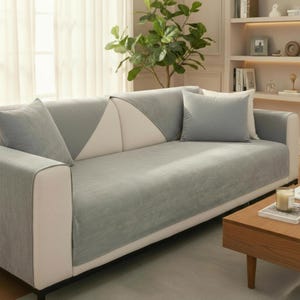 May include: A contemporary sofa featuring a light grey cover and matching cushions. The sofa has a white and grey geometric pattern on the back and armrests. The room is decorated with a plant, a wooden coffee table, and a bookshelf.
