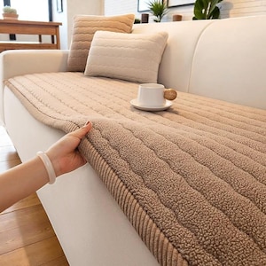 Könnte beinhalten: Eine beige Sofabezug mit einem strukturierten, gerippten Design wird auf einem weißen Sofa gezeigt. Zwei dekorative Kissen und eine weiße Kaffeetasse mit Holzgriff befinden sich ebenfalls auf dem Sofa. Eine Hand hält den Rand der Abdeckung.
