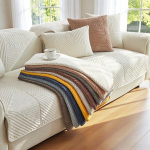 Könnte beinhalten: Ein cremefarbenes Sofa mit mehreren dekorativen Kissen in Creme- und Brauntönen. Ein Stapel gefalteter, gemusterter Überwürfe in verschiedenen Farben, darunter Gelb, Blau und Braun, liegt auf dem Sofa. Eine weiße Teetasse und Untertasse liegen oben auf den Überwürfen.