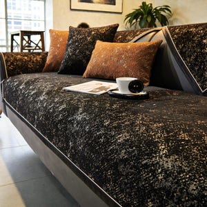 May include: A black and gray patterned sofa with several throw pillows in shades of brown and black. A white coffee cup sits on a black saucer on the sofa. The sofa has a textured, speckled design.