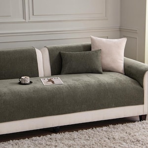 May include: A modern sofa with a green cover and cream-colored sides. Two decorative pillows in green and cream rest on the sofa. A small cup and a magazine are on the sofa. The sofa sits on a light gray rug.