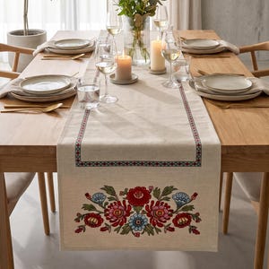 May include: A beige linen table runner featuring a decorative border and a colorful floral embroidery pattern in red, pink, and blue tones. The runner is placed on a wooden dining table set with ceramic plates, gold cutlery, and glassware.