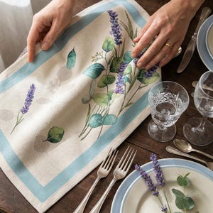 Puede incluir: Un mantel individual de lino beige con un borde azul claro, con un diseño de lavanda y eucalipto. El mantel individual está colocado sobre una mesa de madera con cubiertos, platos y vasos. Primer plano del mantel individual.