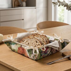 May include: A square fabric bread basket with a colorful autumn landscape print featuring trees and a house. The basket has four corners tied with cream-colored fabric ribbons and contains several slices of rustic sourdough bread.