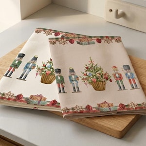 May include: Two cream-colored Christmas-themed kitchen towels with a festive border. The towels feature nutcracker soldiers, Christmas trees, and gift boxes. They are folded and placed on a wooden cutting board, perfect for holiday decor.