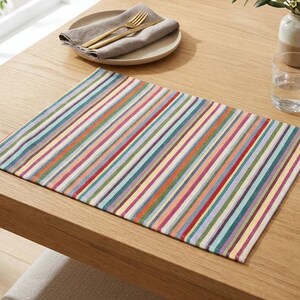 May include: A rectangular placemat featuring a repeating pattern of thin vertical stripes in various colors including red, blue, yellow, green, and cream. The placemat is made of a woven fabric and lies flat on a light-colored wooden surface.