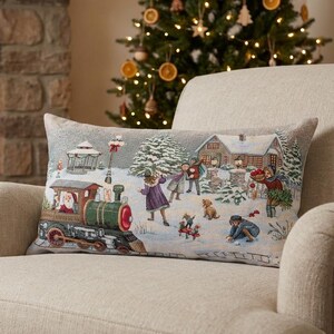 May include: A rectangular cushion with a winter scene depicting a train with Santa, children playing in the snow, and houses. The cushion is on a beige armchair in front of a fireplace and Christmas tree.