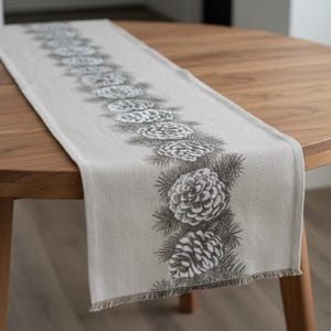 May include: A light gray table runner with a repeating pattern of detailed pine cones and needles in dark gray. The runner is placed on a round wooden table. The runner has a fringed edge.