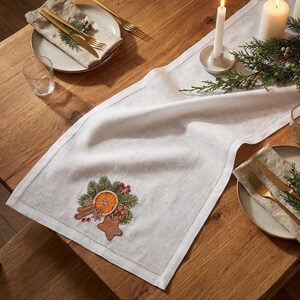 Pode incluir: Uma mesa festiva com um caminho de mesa de linho branco bordado com um design natalino. Talheres dourados, pratos e velas criam um ambiente acolhedor. O caminho de mesa é decorado com uma fatia de laranja e paus de canela.